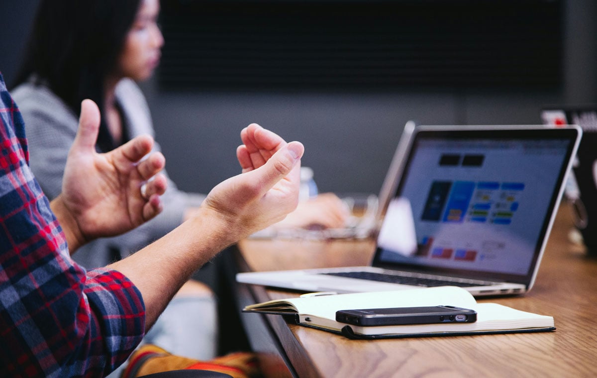 A gesturing pair of hands hover above a work table with an open laptop, notebook and phone.