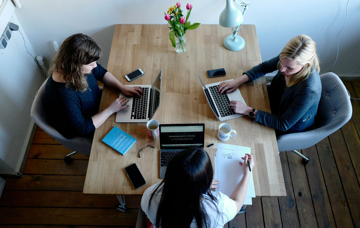 Three women working up a B2B Go To Market Strategy at a wood table with laptops.