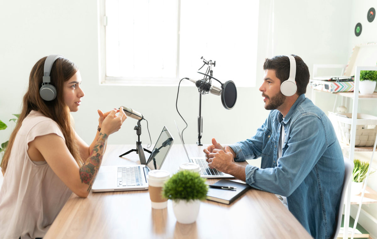 A dark haired woman sits across from a dark haired man at a table with microphone and laptop