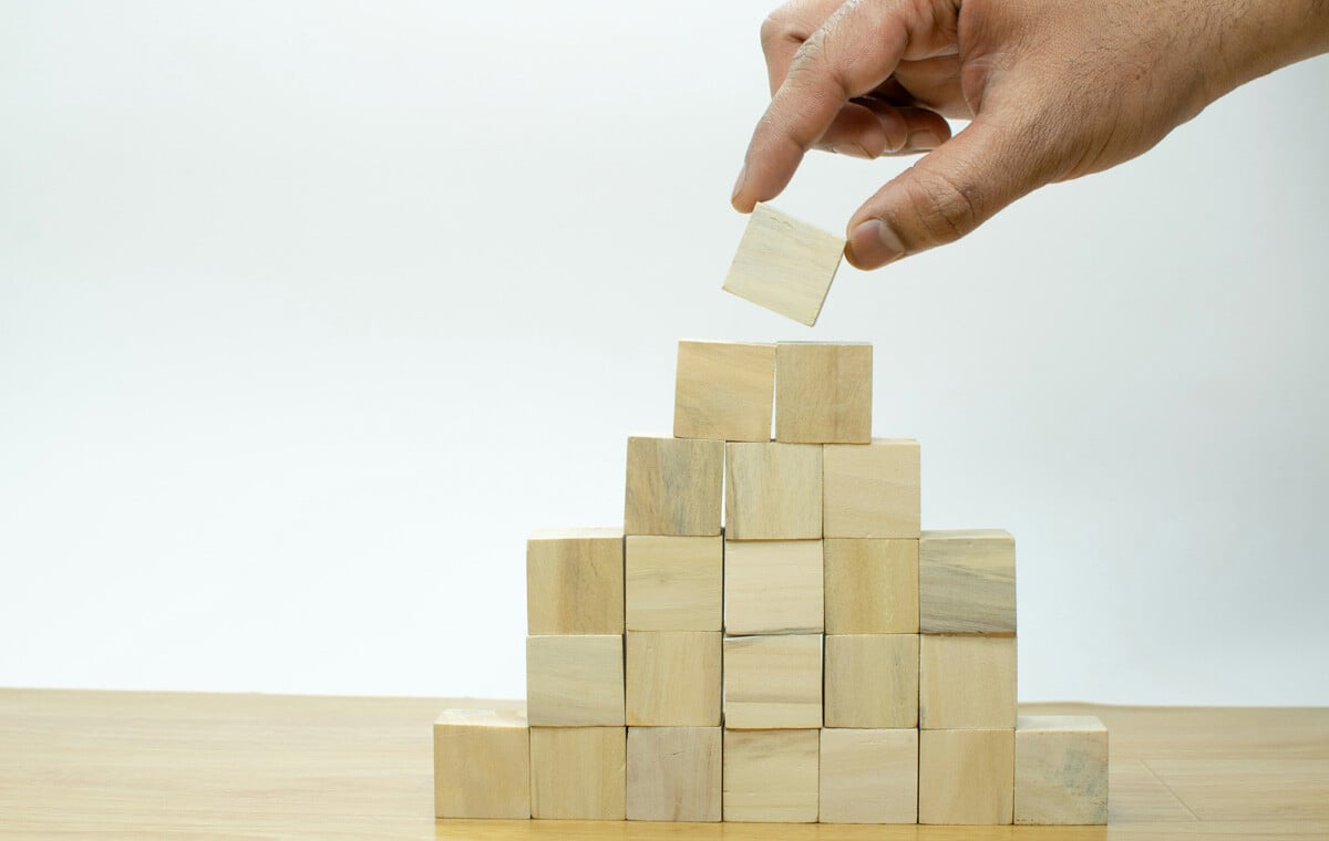 A hand stacking wooden blocks into a pyramid shape on a light wood table.