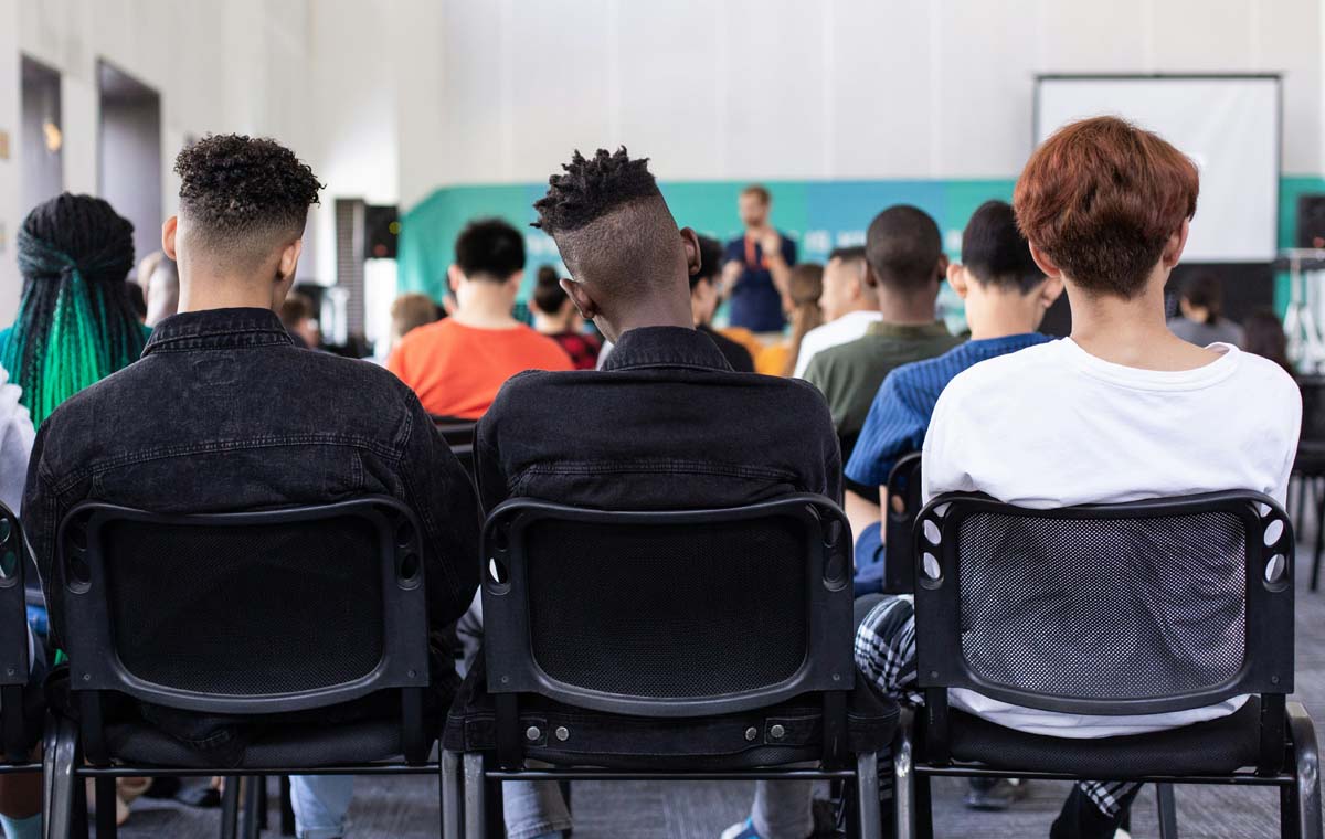 An audience of young people sits with their backs to the camera in a room with a blue wall